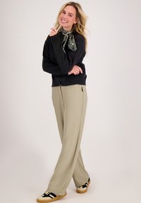 Glunderende vrouw in zwarte trui, groene geprinte sjaal om de nek, beige wijde broek en zwart-witte sneakers op lichte achtergrond.