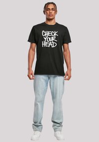 Black cotton t-shirt with a white graphic text "CHECK YOUR HEAD" centered on the front. Styled with light blue jeans and white sneakers.