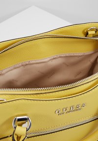 Keltainen nahkainen Guess-laukku, jossa on hopeiset vetoketjut, metallirenkaat ja beige sisävuori, johon on painettu "Guess"-logo.