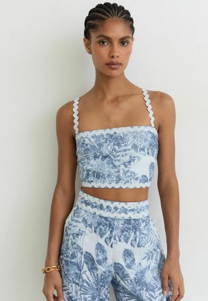 Frau mit geflochtenem Haar, die ein blau-weiß geblümtes Crop-Top mit Zickzack-Trägern und passende hoch taillierte Hosen vor einem schlichten weißen Hintergrund trägt.