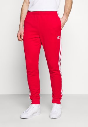Mann trägt rote Adidas-Trainingshose mit weißen Streifen an den Seiten und weiße Turnschuhe vor einfachem Hintergrund.