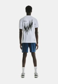 White short-sleeve shirt with black abstract design on the back, paired with blue shorts. Features branding on both shirt and shorts.