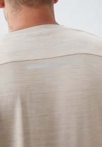 Béžové tričko s hladkým povrchom, s okrúhlym výstrihom a jemným logom potlačeným pri šve na pleci. Strih prispôsobený na telo, minimalistický dizajn.