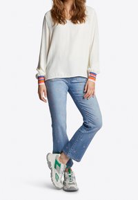 Hvid langærmet bluse med stribede manchetter, parret med lyseblå jeans med blomsterbroderi og hvide sneakers med grønne accenter.