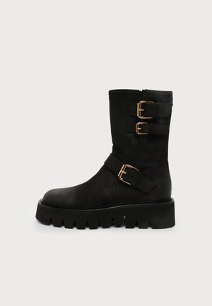 Schwarzer Stiefel bis zur Mitte der Wade mit dicker Profilsohle und drei goldenen Schnallenriemen an der Seite, vor einem schlichten hellen Hintergrund dargestellt.