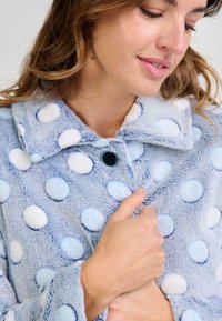 Abrigo de piel sintética en azul claro con grandes lunares texturizados en blanco y azul claro. Presenta un cuello y un cierre de botón negro.