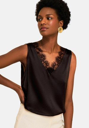 Frau mit kurzen lockigen Haaren trägt ein schwarzes ärmelloses Satintop mit Spitzenbesatz und großen goldenen runden Ohrringen, posiert mit der Hand in der Hüfte.