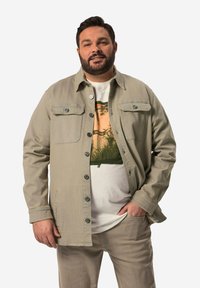 Khaki nappatakki, jossa on kaksi rintataskua, käytetään graafisen t-paidan päällä. Siinä on rento istuvuus ja sileä pinta. Vaaleat housut.