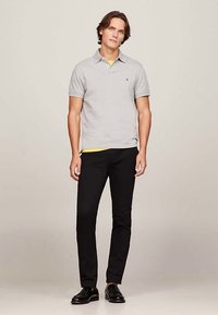 Mladý muž stojící, oblečený v světle šedém polo tričku, černých kalhotách, černých botách a žlutém tričku, které je vidět na lemu.