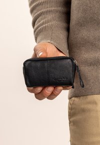 Czarny skórzany portfel na zamek, o gładkiej teksturze i subtelnych detalach przeszycia. Posiada małą pętlę do mocowania. Trzymany w ręku dla skali.