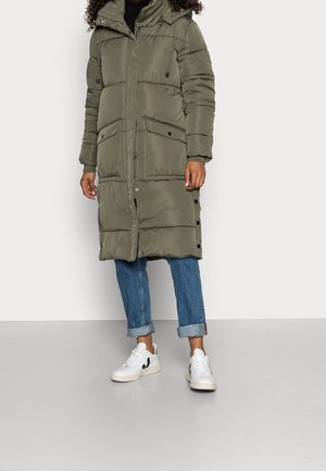 Personne portant un long manteau matelassé vert olive avec de grandes poches avant, un jean bleu retroussé et des baskets blanches sur un fond clair.