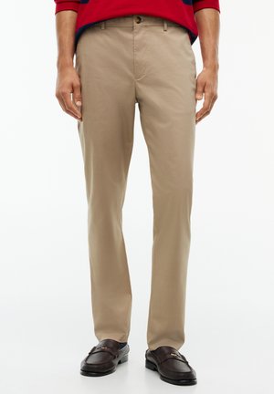Mann trägt beige gerade geschnittene Hose, dunkelbraune Loafers und ein rotes Langarmshirt mit marineblauen Streifen.