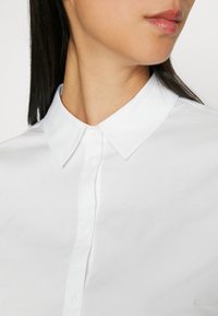 Chemise en coton blanc avec col pointu, boutonnage frontal et texture lisse mettant en valeur des lignes épurées et un design minimaliste.