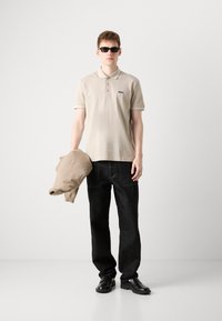 Beige Poloshirt mit Sportkragen, dunkle Jeans, hält eine hellbraune Jacke, trägt schwarze Sonnenbrillen und schwarze Schuhe, einfarbiger Hintergrund.