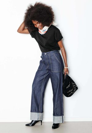 Femme aux cheveux bouclés portant un haut noir, un jean large retroussé, des bottines à talons noires, une écharpe, et tenant un sac à main noir clouté contre un mur blanc.