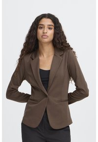 Brun skräddarsydd kavaj med nedvikt slag, två framfickor och en figurnära silhuett. Tillverkad av ett slätt tyg i enfärgat.