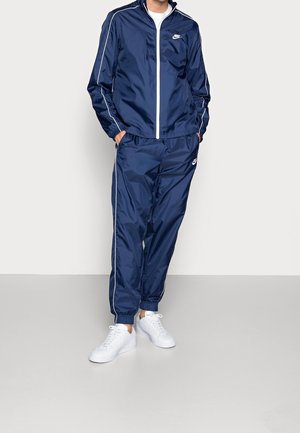 Personne portant un survêtement Nike bleu marine avec des liserés blancs et des baskets blanches, debout les mains dans les poches devant un fond uni.