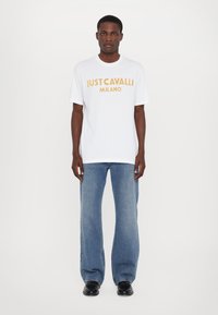 Bílý bavlněný tričko s výrazným zlatým nápisem "JUST CAVALLI MILANO", doplněné modrými džínami a černými botami.