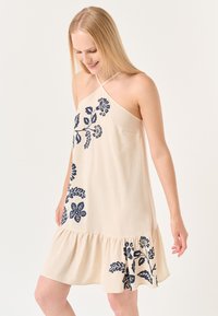 Beiges ärmelloses Kleid mit marineblauem Floralprint, schwingendem Rüschensaum und überkreuzten Trägern. Weicher Stoff und leichtes Material.