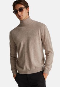 Hellbrauner, gestrickter Rollkragenpullover mit langen Ärmeln und gerippten Bündchen, kombiniert mit schwarzen Hosen. Glatte Textur, lässiges Design.