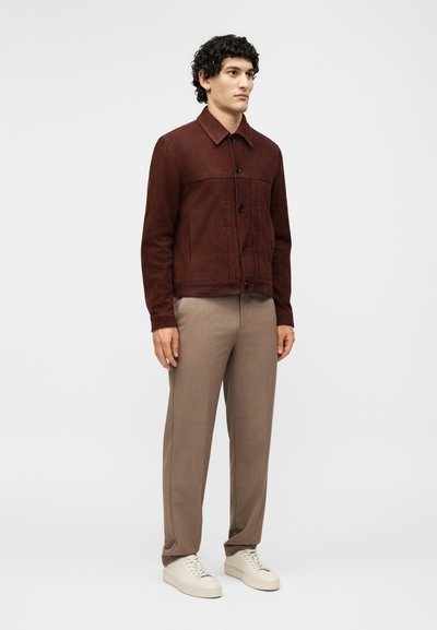 Άνδρας που στέκεται φορώντας ένα σκούρο καφέ σακάκι με κουμπιά, μπεζ παντελόνια και λευκά αθλητικά παπούτσια σε απλό ανοιχτό φόντο.