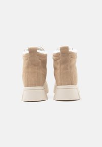 Botas de tornozelo em camurça bege com forro de lã branca, apresentando uma sola de borracha creme e detalhes costurados. A vista traseira mostra um design arredondado.