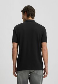 Polo nero a maniche corte, con colletto classico e texture liscia, abbinato a jeans grigi. Visto da dietro.