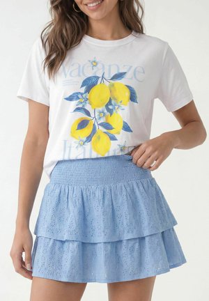 Kobieta ubrana w biały t-shirt z żółtymi cytrynami i niebieskim kwiatowym wzorem, w połączeniu z jasnoniebieską warstwową spódnicą z ażurowym wzorem.