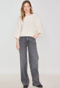 Pull en tricot crème surdimensionné avec un col bateau, associé à un jean gris à taille haute et à pattes d'éléphant, ainsi qu'à des chaussures noires. Design simple et texturé.