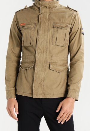 Veste utilitaire pour homme couleur kaki avec plusieurs poches frontales, épaulettes aux manches et col montant, portée avec un pantalon noir.