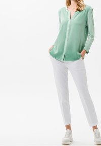 Camicia verde chiaro a maniche lunghe con scollatura a V e chiusura con bottoni, abbinata a pantaloni bianchi sartoriali e sneakers bianche. Texture liscia, design casual.