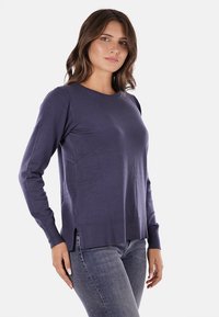 Maglione lungo viola scuro a maniche lunghe con collo rotondo, polsini e orlo a costine, dotato di fessure laterali per facilitare il movimento. Texture liscia.