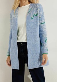 Hellblauer, gestrickter Cardigan mit grünem Blumenmuster, offenem Design, gerippten Manschetten und einer weichen Textur. Kombiniert mit einem weißen Oberteil und dunklen Jeans.