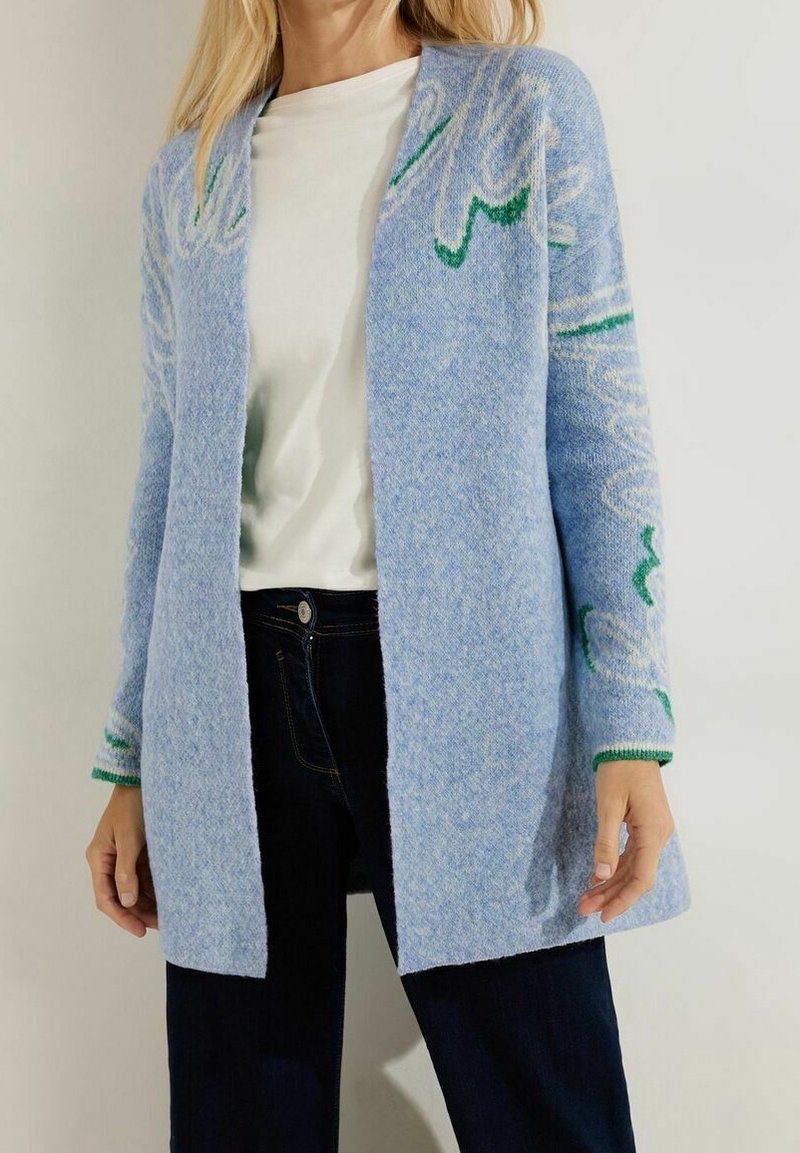 Hellblauer, gestrickter Cardigan mit grünem Blumenmuster, offenem Design, gerippten Manschetten und einer weichen Textur. Kombiniert mit einem weißen Oberteil und dunklen Jeans.