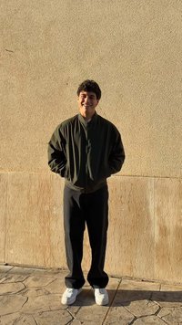 Bărbat îmbrăcat cu o jachetă bomber verde închis, pantaloni negri și adidași albi, stând în fața unui perete texturat bej, zâmbind.