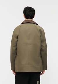 Άτομο που φοράει μπουφάν ελιάς με καφέ γιακά, γυρισμένο προς τα πίσω, με απλό φωτεινό φόντο.