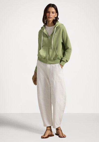 Grønn hettejakke med glidelås, snorer, frontlommer og logo. Kombinert med løse hvite bukser og flate sandaler. Enkelt bakgrunn.