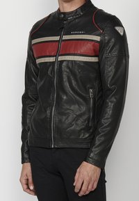 Giacca di pelle nera con strisce orizzontali rosse e beige, chiusura frontale con zip e patch con marchio sulla manica superiore sinistra, indossata da una persona in piedi.