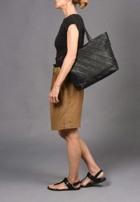 Bolso tote negro de tejido con correas largas, combinado con una blusa negra de manga corta y una falda marrón hasta la rodilla, llevado con sandalias negras.