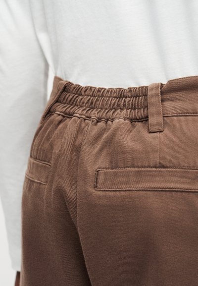 Καφέ παντελόνι με ελαστική μέση με πιέτες και θηλιές ζώνης που φοριέται με λευκό μπλουζάκι μακρυμάνικο, εστίαση στη μέση του πίσω μέρους και τις λεπτομέρειες της τσέπης.