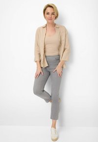 Beige linnen shirt over een beige tanktop, gecombineerd met lichtgrijze broek en crèmekleurige sneakers, tegen een effen witte achtergrond.