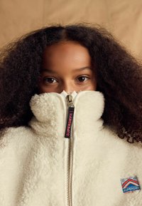 Veste zippée en polaire blanche avec un col haut, une fermeture éclair en métal et une étiquette de logo noire. Présente une surface texturée et une pièce colorée sur le côté.
