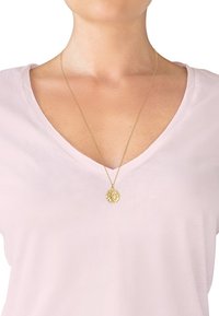 Cou et haut de poitrine d'une personne portant un haut à col en V rose pâle et un collier en chaîne dorée avec un pendentif rond présentant un profil en relief.
