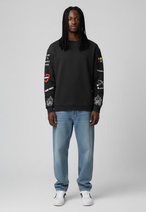 Schwarzer Hoodie mit bedruckten Designs an den Ärmeln, kombiniert mit hellblauen Jeans und weißen Sneakers. Besitzt einen lockeren Schnitt und einen Rundhalsausschnitt.