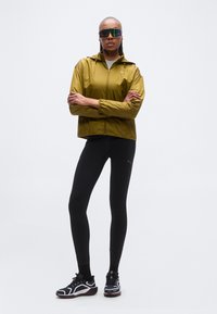 Chaqueta verde ligera con capucha, combinada con mallas negras y zapatillas deportivas. Las gafas de sol tienen lentes reflectantes de colores.
