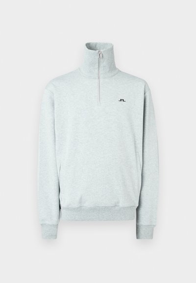 Gaišs pelēks ceturtdaļas rāvējslēdzēja pulovers ar augstu apkakli, garām piedurknēm, ribotiem aprocēm un apakšmalu, kā arī mazu melnu logotipu kreisajā krūtī.