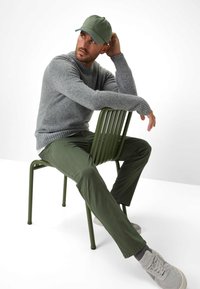 Grijze gebreide trui, groene pet, olijfgroene broek en lichtgrijze sneakers, zittend op een groene stoel met horizontale latten, in een minimalistische witte achtergrond.