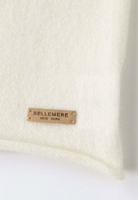 Tessuto bianco lavorato a maglia con una superficie liscia e un bordo arrotondato. Un'etichetta rettangolare in pelle marrone con la scritta "BELLEMERE NEW YORK" è attaccata.