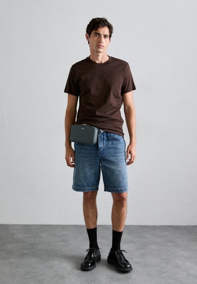 Ung mann iført mørkebrun t-skjorte, denimshorts, svarte sokker og polerte svarte sko, stående mot en ensfarget lys veggbakgrunn.