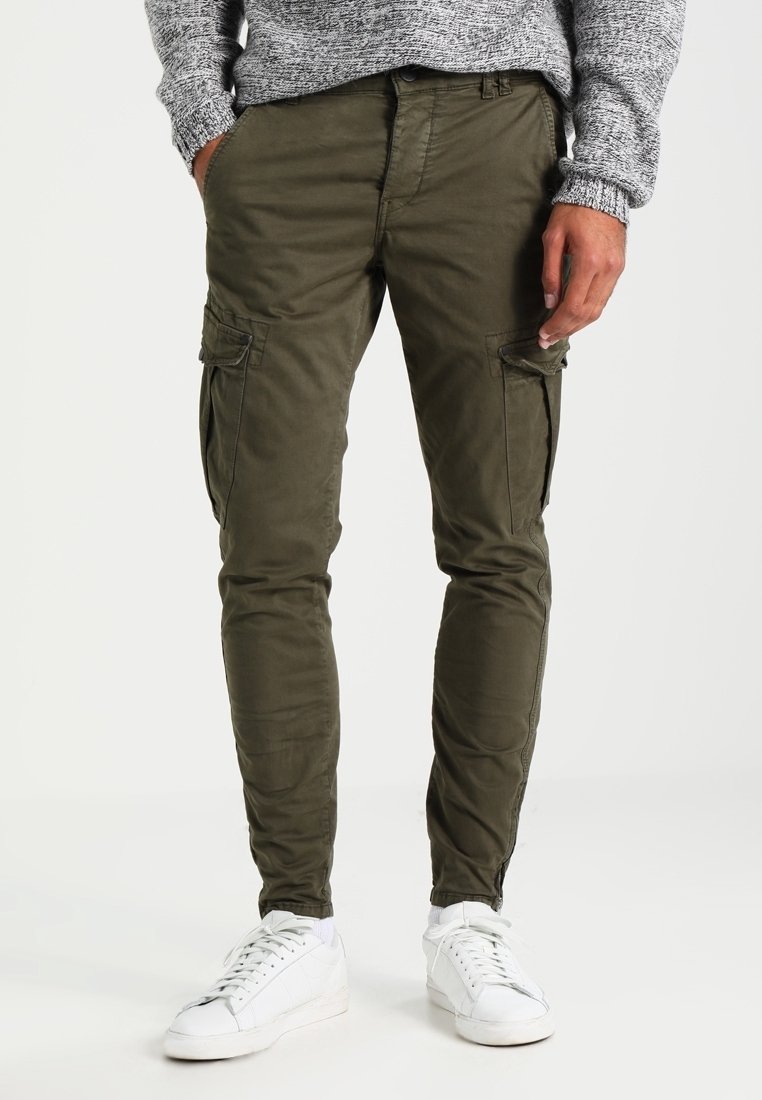 Man in olijfgroene cargobroek, grijze trui en witte sneakers, staande met handen ontspannen langs zijn zijde.